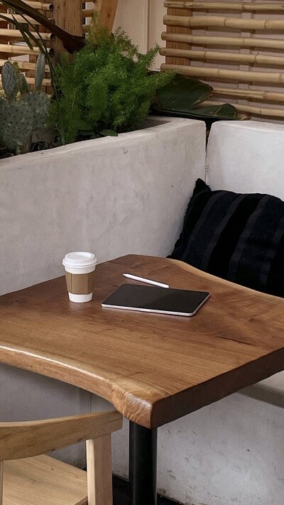 A cozy wooden table with a black notebook, pen, and takeaway coffee. Green plants and a black cushion on a bench add a natural, relaxed feel.