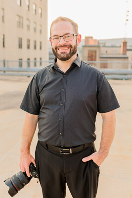 White Male Scranton Wedding photographer Eric Boylan wearing a holdfast harness and black vest  holding a Canon R6 while sitting on a stool against a beige backdrop