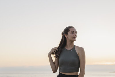 Katie looks off into the distance with her hand on her hair in front of the ocean.