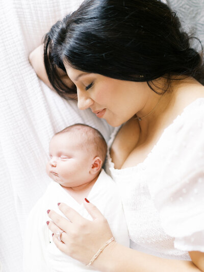Brunette mother in white dress lies on bed snuggling her swaddled newborn baby boy