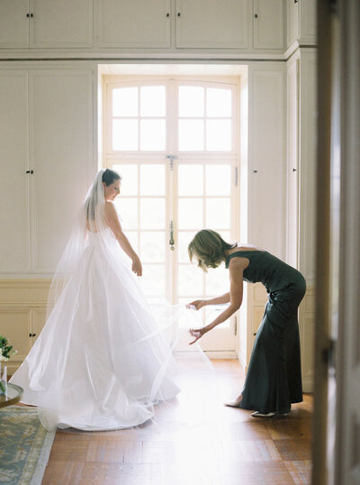 Rhode Island Wedding Photographers | A bride in a white gown and veil stands near a bright window. A woman in a dark dress adjusts the gown's train.