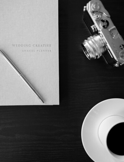 linen creative journal with gold foil lettering, gold pen, vintage camera, and cup of coffee on a black table