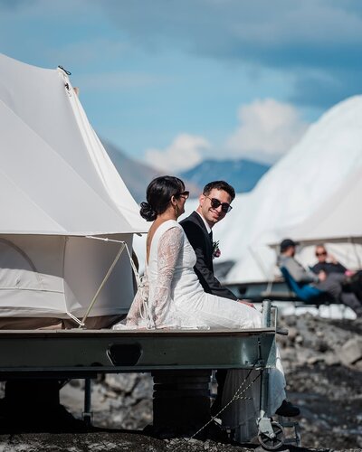 A white canvas glamping bell tent stands beautifully atop a platform that sits on the surface of the glacier.  on the edge of the platform, a bride in a beauitufl lace dress and groom rest comfortably, smiling and lauging.  The groom is looking towards the camera, wearing sunglasses with a huge smile during their adventure luxe elopement with glamping on a glacier in alaska. 