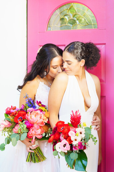 Two brides at their wedding standing with bouquets in front of a pink door at Rancho Las Lomas 