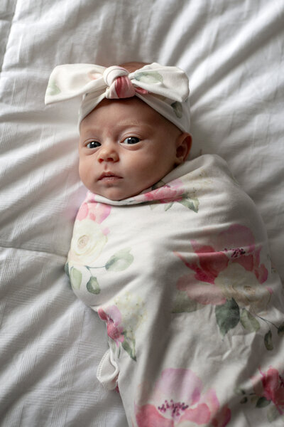 Newborn girl wrapped in white floral wrap, lying on white comforter. 