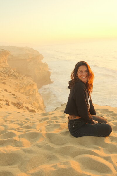 A woman sits quietly beside a cliff’s edge, the ocean stretching before her as the sun dips below the horizon in a wash of golden light.