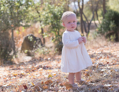 Baby in a white dress stands in a sunlit field, gazing curiously at the camera. A timeless baby milestone session by Moments in Light Photography.