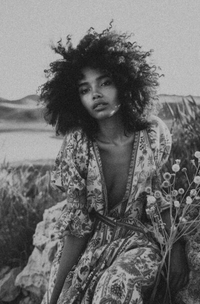 A woman with curly hair sits outdoors in nature, wearing a patterned dress with a deep neckline. Captured in black and white, this photo reflects the timeless aesthetic often embraced by Sweet Pea Ink Creative’s approach to brand design.