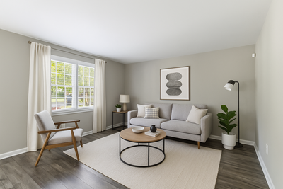 Contemporary living room with a gray sofa, modern artwork, large windows with white curtains, and hardwood floors.
