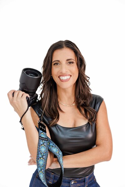 Smiling woman holding a camera, wearing a black top and jeans, during a brand photo session for Branding by Jo.