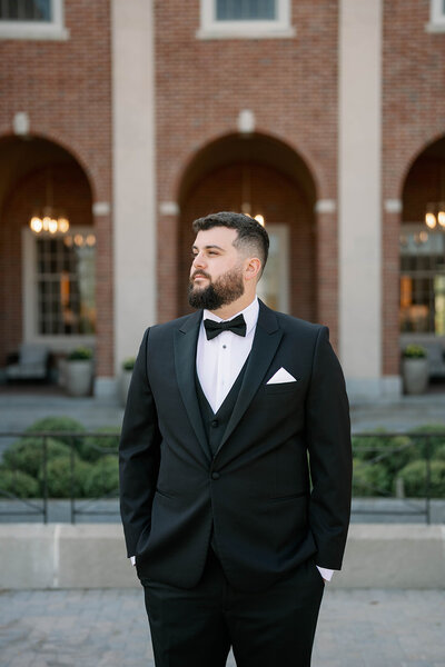 Editorial portrait of a groom in a black tuxedo standing outside a brick venue in Michigan, taken in a timeless and true-to-color wedding photography style.