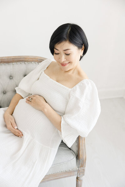 Pregnant woman in white flowing dress sits on a grey settee and smiles while embracing her pregnant belly during maternity photography session