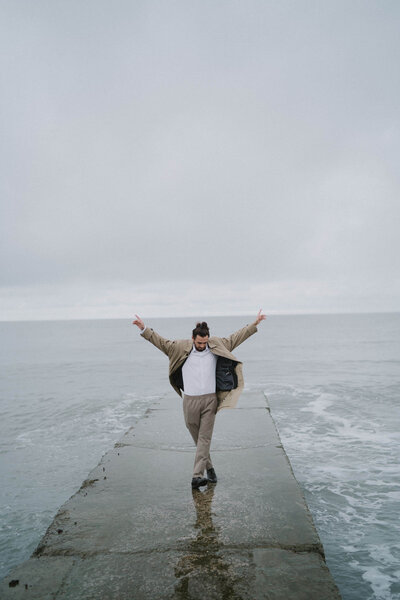 Mann auf einer Boots-Slipanlage am Ostsee-Strand, die Beine über Kreuz und beide Arme triumphierend nach oben streckend: Selbstbewusstsein und Erfolgsgefühle erleben.