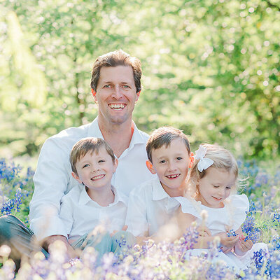 Father holding three children in his lap in a field of bluebonnets.