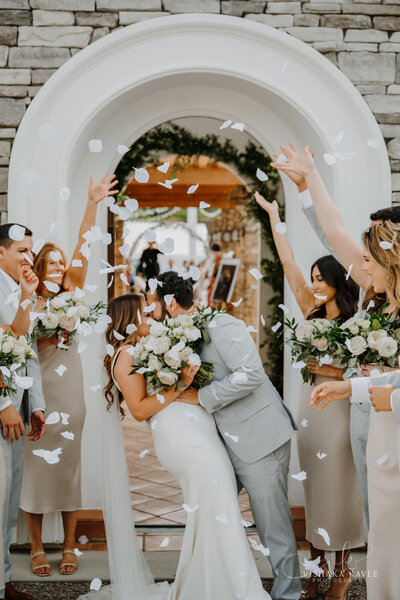 bride and groom kissing while bridal party throws flower petals all around them
