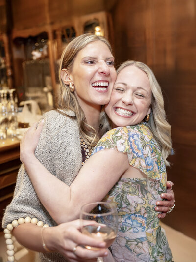 Two women smiling and hugging warmly in a warmly lit room. One holds a glass of wine. They appear joyful and the setting suggests a social event or celebration, with blurred background elements that include shelves and glassware.