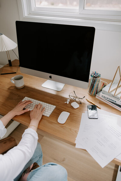 Website copywriter Erika Fitzgerald typing on an iMac during the research phase of website copywriting, with strategy notes and papers on the desk