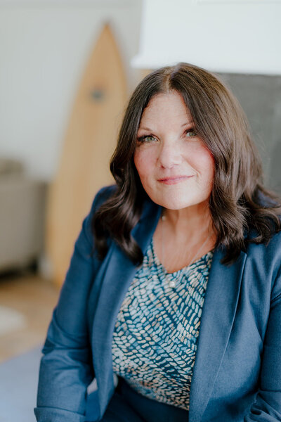 Close-up portrait of Susie Schumacher seated indoors, wearing a navy blazer and patterned blouse, with soft natural light highlighting her face and a surfboard visible in the background – Susie Schumacher Life Coach