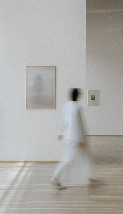 blurry capture of woman walking away in white outfit in gallery with artwork on walls