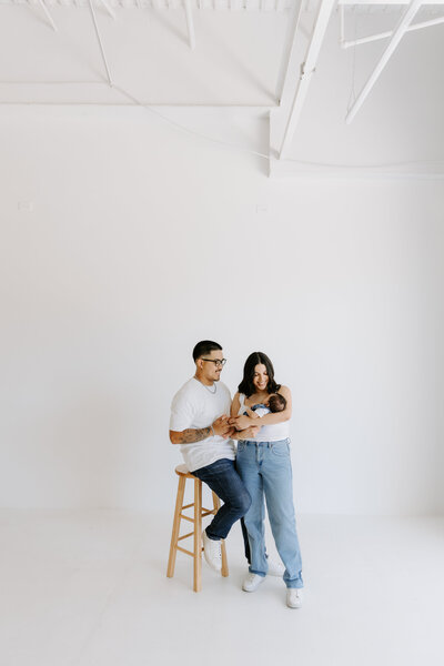 A couple sits in a minimalist white studio holding their newborn baby, capturing a quiet and loving family moment.