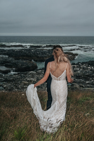 Romantic wedding moment by the Pacific Northwest ocean, featuring a flowing white dress caught in the wind against dramatic dark rocky cliffs, captured on the Oregon coast by a Portland wedding photographer.
