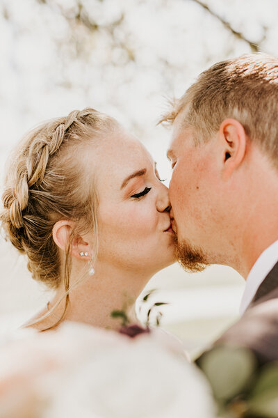 A blonde bride with a braid along the crown of her head kissing her groom with bouquet in foreground