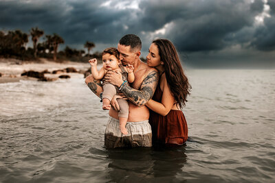 Family Photographer, a father holds his baby as mom looks on, they stand in the quiet ocean waters