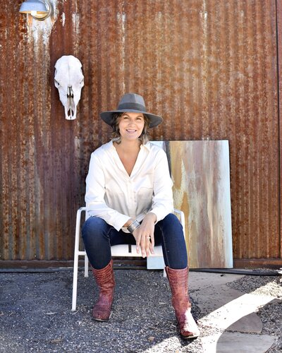 woman smiling while sitting on chair in jeans and white shirt and hat with painting behind her