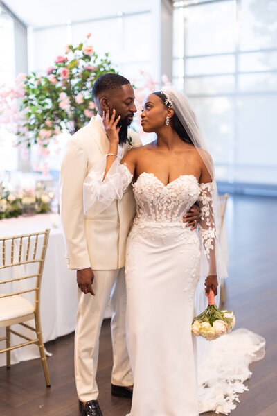 Groom standing beside bride on her left side, holding her by her waist as she places her hand on his face and looks into his eyes.