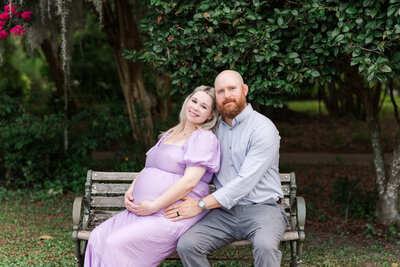 mom and dad snuggling while holding baby bump seating at the park