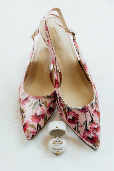 A bright pink pair of floral shoes with an engagement ring in a vintage-style ring box.