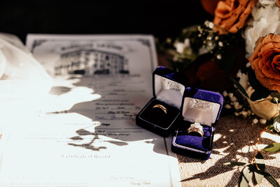 The bride and groom's rings in their blue velvet boxes set against a background of the pew, the bride's bouquet, and the marriage license