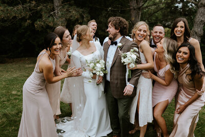 A bride and groom stand smiling, surrounded by a joyful group of bridesmaids and groomsmen for their estate wedding at Newmarket, all dressed in formal attire, outdoors with trees in the background. Captured by Jennifer van Son, a talented Muskoka wedding photographer, this moment reflects pure happiness and natural beauty.