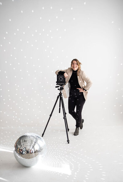 A person smiling as they lean one arm on an old school camera with a disco ball on the floor in front.