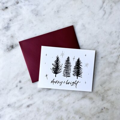 Card with an illustration of pine trees and words that read "Merry + Bright" in cursive script