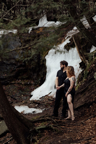 bride and groom both dressed in all black stand on a path that is deep in the woods surrounded with thick ice from a frozen waterfall. the bride and groom are looking out over the edge of the trail 