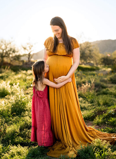 Beautiful mom-to-be in yellow gown hugging older daughter. Captured by Cactus & Pine Photography