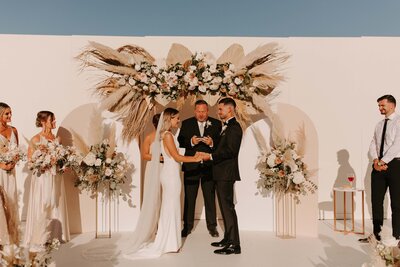 Bride & Groom in Front of Boho Ceremony Backdrop - Bre & Chris | Converted Basketball Court Wedding – Featured in Brides Magazine