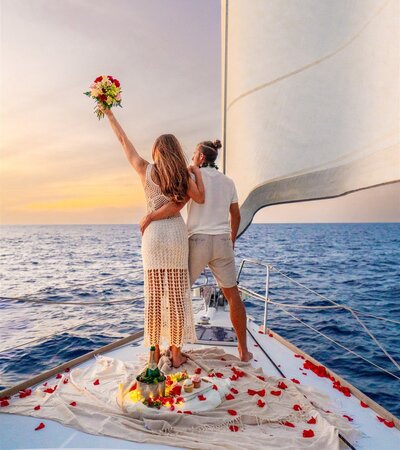Couple standing on the front of a sailboat at sunset, celebrating with rose petals and champagne as they look out over the ocean in Hawaii.