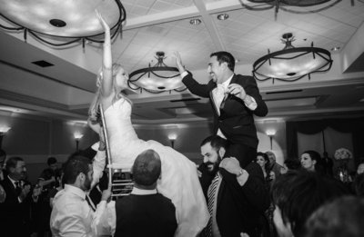 Bride and groom hoisted into the air on chairs at their Jewish wedding reception, in black and white