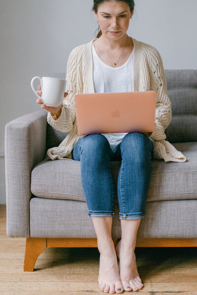 Woman sits on couch working at laptop and holding coffee mug