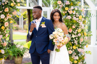 Bride and groom photograph.  Bride with large blue eyes and a white fitted wedding gown looking off in the distance towards the left.  Groom is lovingly looking at his Bride.  Groom holding bride at waist.  Bride holding on the grooms upper arms in an embrace