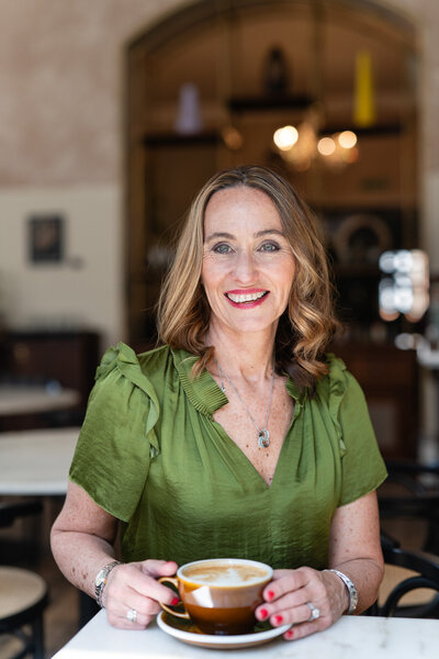 woman sitting at a desk with a cup of coffee wearing a green shirt and smiling to a camera