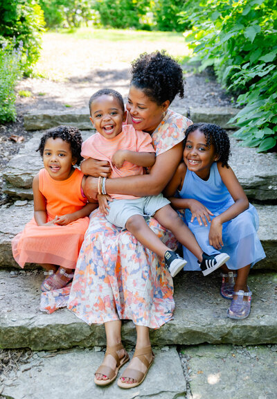 Smiling woman in a colorful white, orange, and blue patterned dress poses for a family portrait, surrounded by two cheerful young girls, with a happy little boy sitting on her lap.