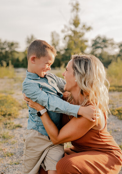 mom in burnt orange dress squatting down to hold son in blue jean button up and khaki shorts holding her back, they're looking at each other.