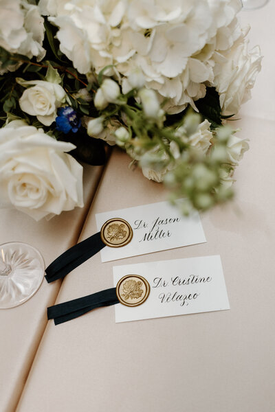 Wedding place cards with gold wax seals and black ribbon, styled next to a white floral bouquet.