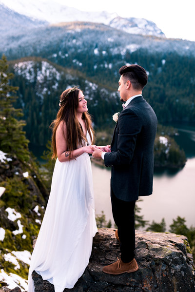 couple holding hands saying vows atop a snowy mountain with lake in distance