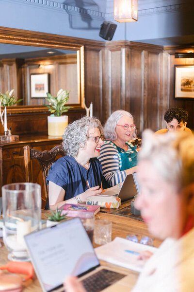 In a cosy, wood-panelled room, a curly grey-haired person in a navy dress chats and smiles with a person in teal dungarees, striped sleeved top and pink glasses; another person listens nearby. Laptops, notebooks, and plants sit on the long table, with a blurred figure in the foreground.