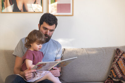 Livsstilsfoto av far som sitter på en grå sofa og leser bok til datteren som sitter på fanget hans.