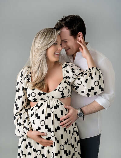 Intimate studio maternity portrait by Stamford CT photographer featuring expecting couple in coordinated black and white patterned dress and white shirt against neutral backdrop